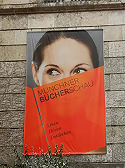 Das Poster der Bücherschau mit großem Frauenkopf der hinter einem roten Buch mit Ausstellungsangaben, deren Augen verschmitzt nach links blicken