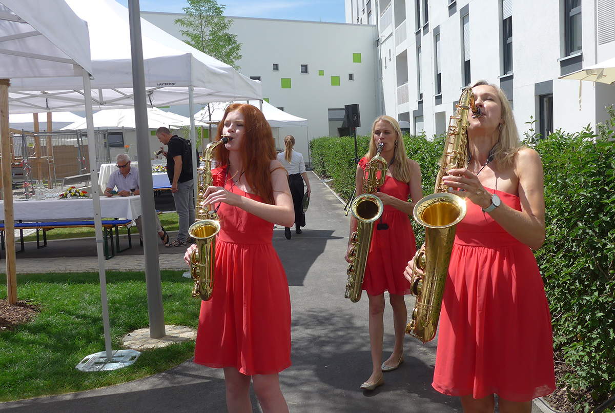 3 Musikerinnen am Saxophon in roten Kleidern gehen musizierend durch den Innenhof von WG 5 bei srahlendem Sommerwetter