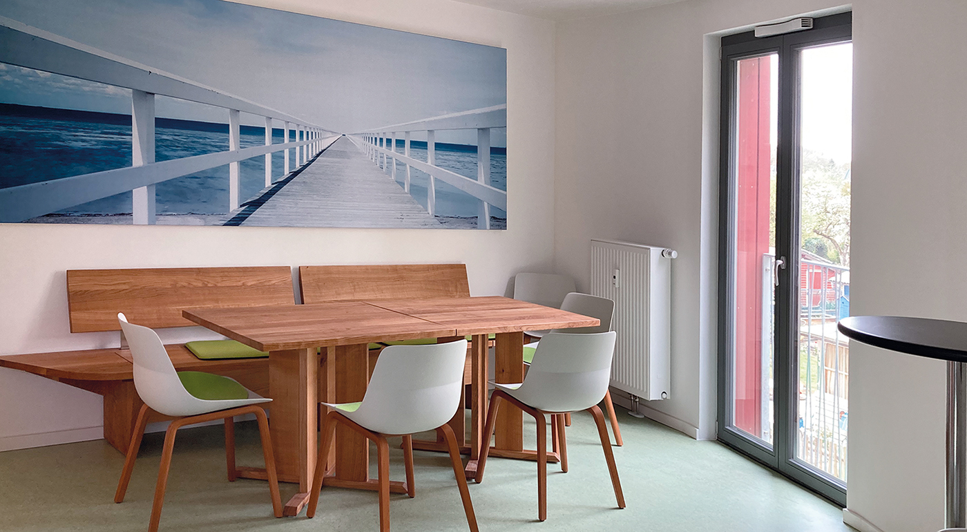 wg6: Blick in den Gemeinschaftsraums zum großen Tisch, rechts ein großes Fenster mit französischem Balkon, über dem Tisch ein Bild von einem langen Steg am Meer